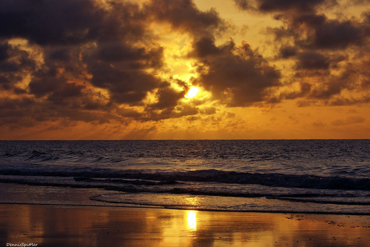 Dramatic golden ocean sunset with sun breaking through dark storm clouds reflecting on wet sand